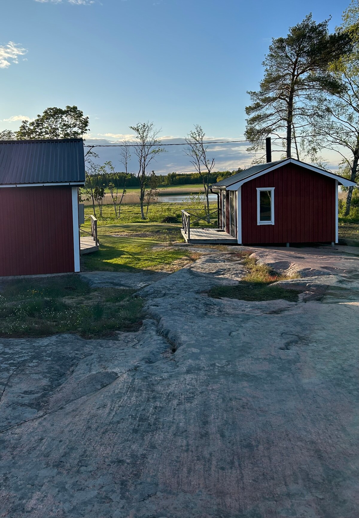 A serene countryside scene depicts two red wooden buildings surrounded by a natural landscape. Green grass and sparse trees frame the area, with a view of the distant fields. Sunlight enhances the tranquil atmosphere, highlighting the rustic charm of the setting.