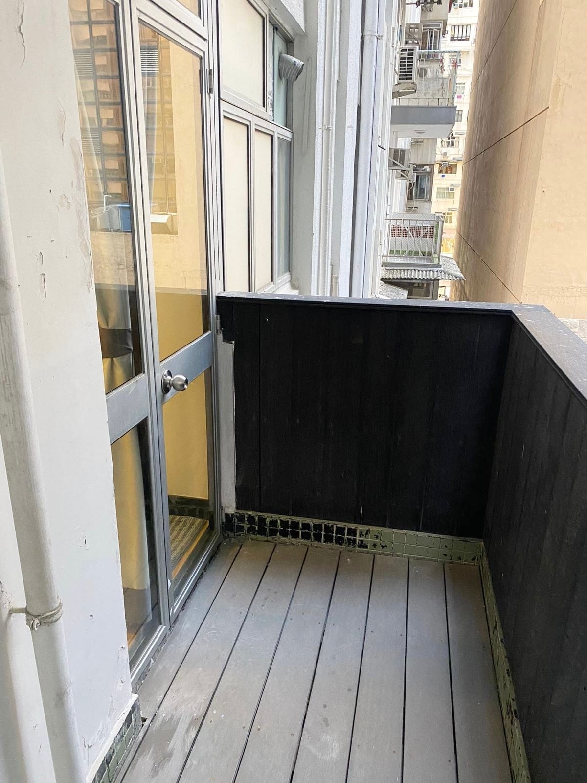 A narrow balcony is visible, featuring wooden flooring and a partial black railing. Glass doors lead into the unit, allowing natural light to enter. Surrounding walls of neighboring buildings create a compact outdoor space.