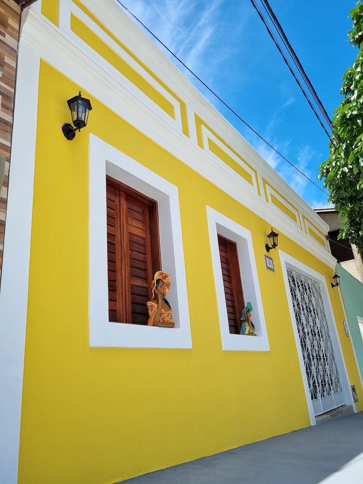 The exterior of a colonial-style house is shown, featuring bright yellow walls contrasted by white trim. Two wooden windows, adorned with decorative figures, are positioned along the facade. Soft blue skies and greenery are visible in the background, enhancing the inviting appearance of the property.