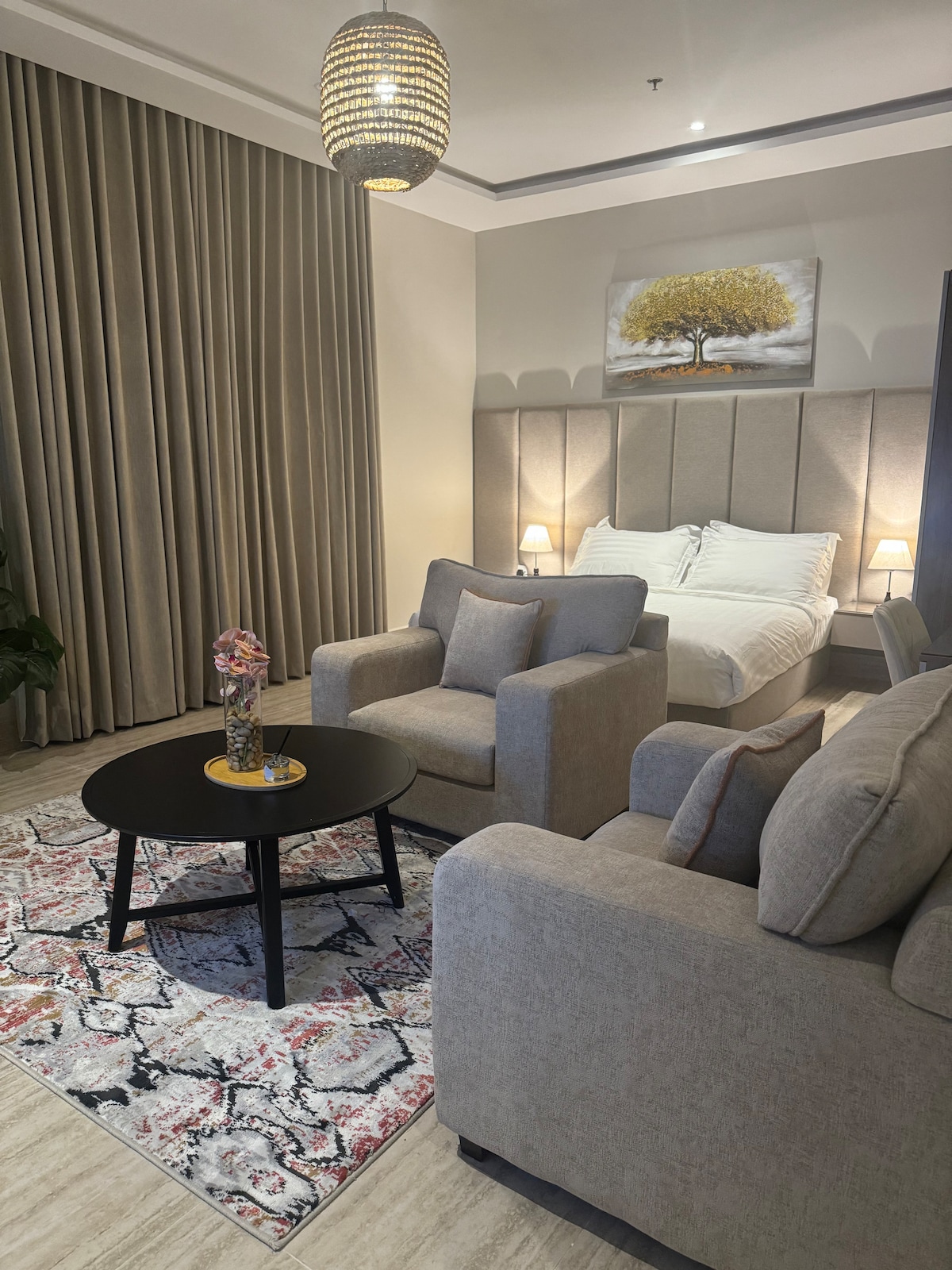 A calm living area features two light gray armchairs positioned around a circular table. A colorful vase holds flowers on the table. A spacious bed is visible in the background, framed by soft, neutral curtains and complemented by a decorative wall art piece above.