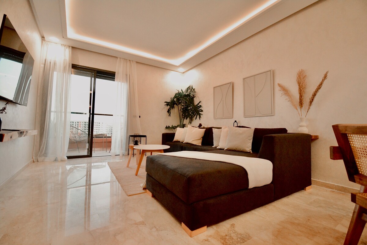 The living room area features a spacious dark sofa with soft cushions, complemented by a light-colored area rug. A flat-screen TV is mounted on the wall, and natural light is welcomed through sliding glass doors, leading to a sunny balcony. Decor elements include plants and minimalist art.