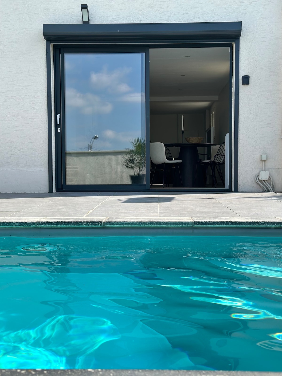 The image captures a serene view of a private pool, with clear turquoise water reflecting the sky. A glass door leads into a tastefully furnished interior, where light and openness are emphasized. The outdoor space features a smooth tiled surface alongside the pool.
