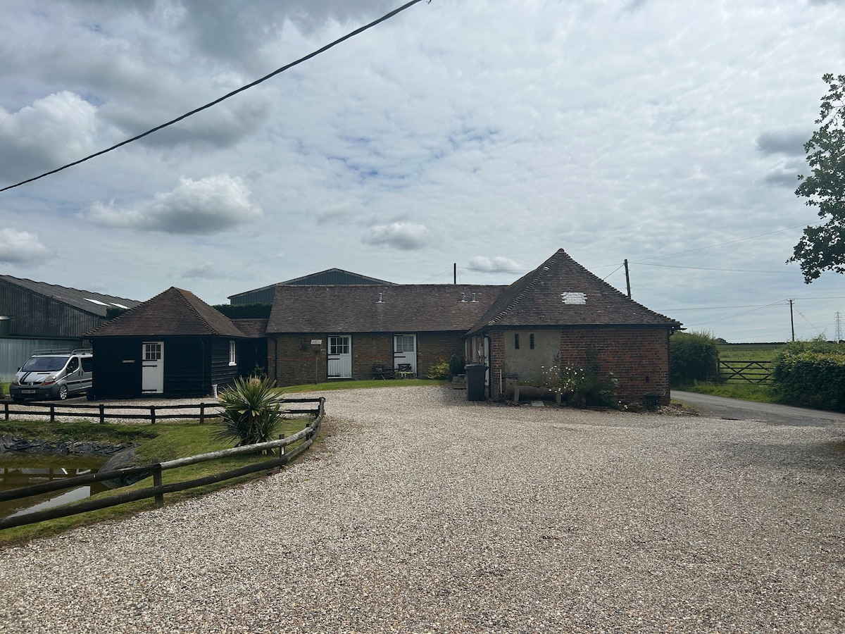 A gravel driveway leads to a charming historic property featuring a main building and two rounded-roof structures. Lush green fields are visible in the background, and scattered plants add a touch of greenery. The sky is partly cloudy, creating a gentle light across the scene.