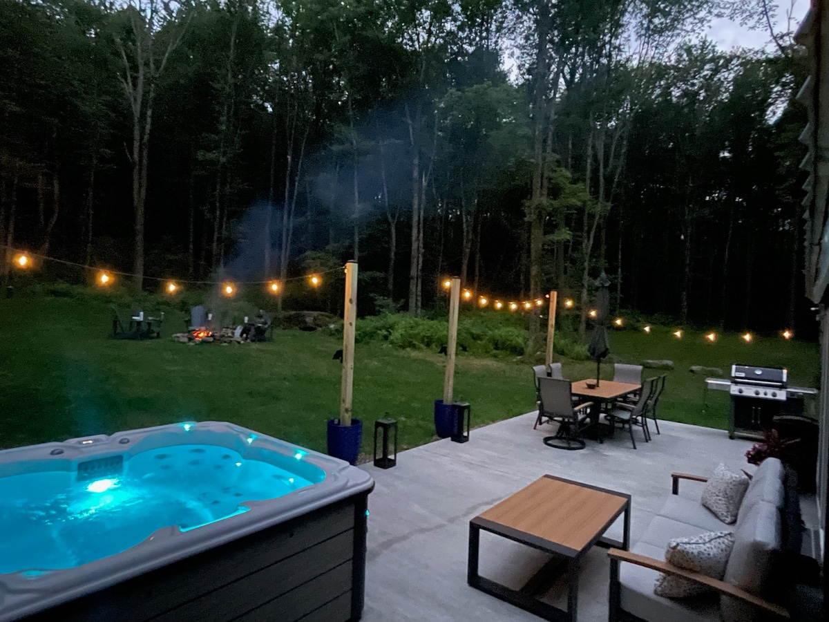 A spacious outdoor area contains a hot tub with soft blue lighting, inviting relaxation. String lights hang overhead, illuminating a dining table and chairs. In the background, fire pit seating is set among the trees, enhancing the serene, wooded ambiance.