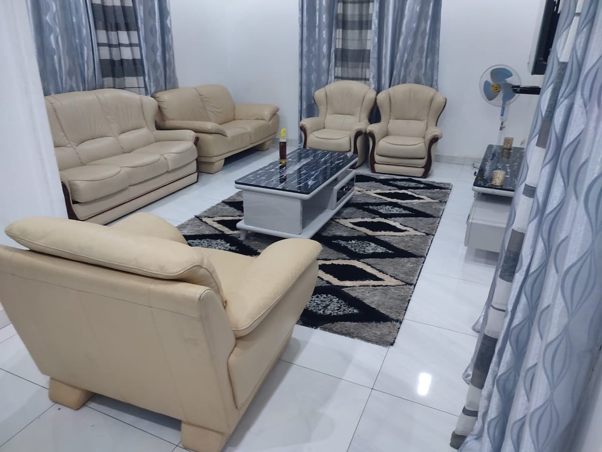 A living area features light-colored leather sofas complemented by patterned armchairs. A coffee table sits centrally on a geometric patterned rug, enhancing the space. The room is adorned with sheer curtains that allow natural light to filter in, creating a calming atmosphere.
