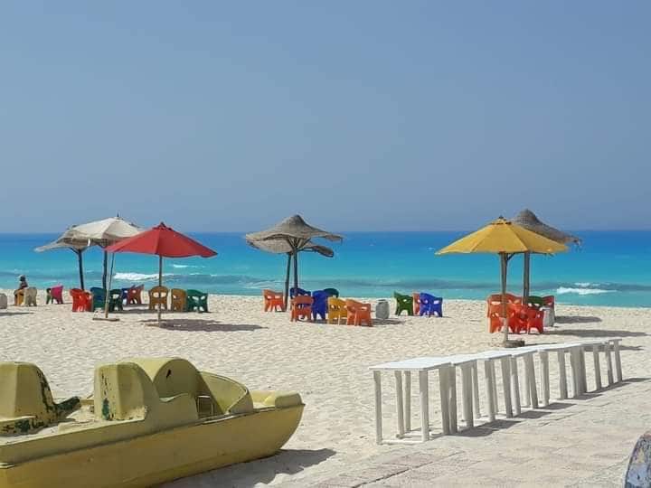 A sandy beach is dotted with colorful chairs under umbrellas, creating inviting spaces for relaxation. Calm waves gently lap at the shore, while a small group of chairs is arranged nearby, offering a comfortable spot to enjoy the coastal views.