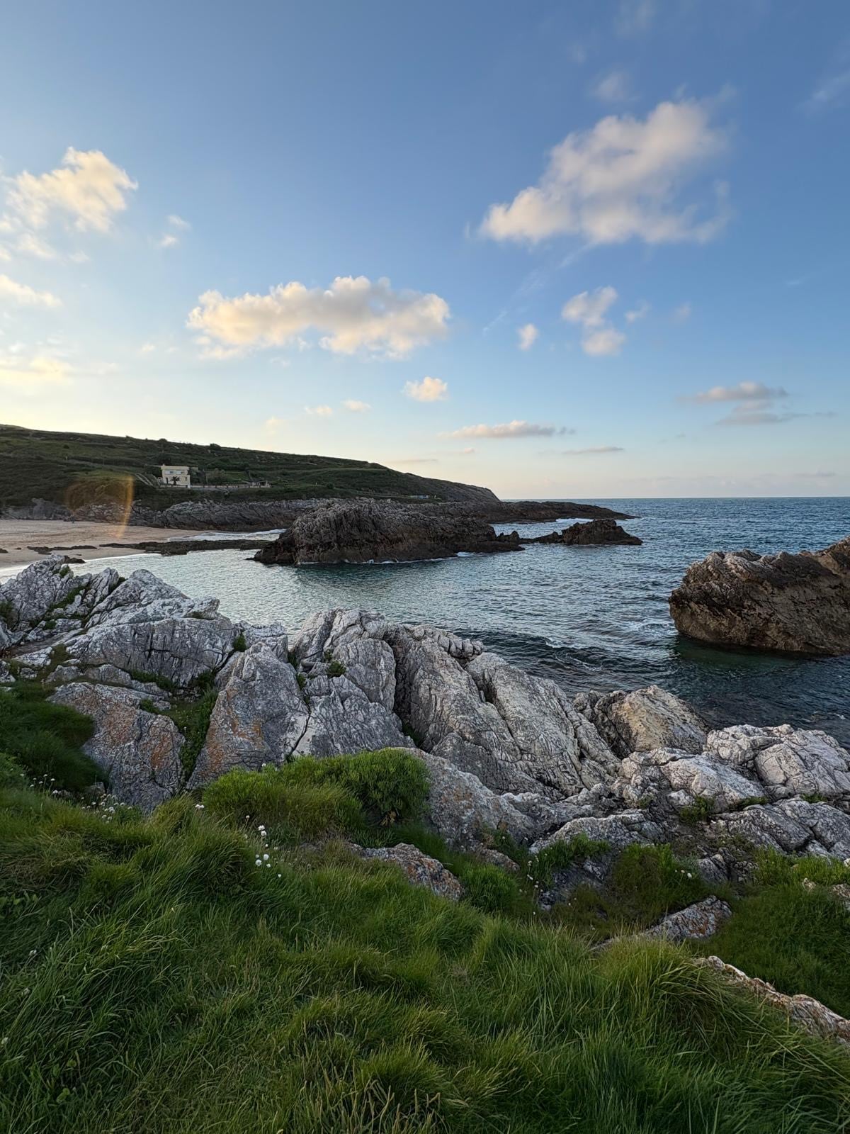 A rocky shoreline is visible, featuring uneven granite boulders covered with patches of green grass. The water is calm, reflecting the soft light of the sky. Gentle waves lap against the shore, while rolling hills are observed in the background under a partly cloudy sky.
