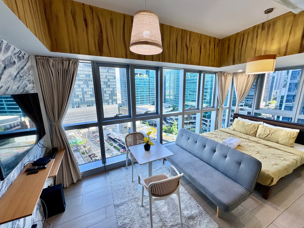 A sunlit living area is showcased with large windows, offering views of the cityscape. A cozy bed is positioned adjacent to a modern seating arrangement, featuring a sofa and a small dining table. Neutral tones and a textured accent wall add warmth to the space.