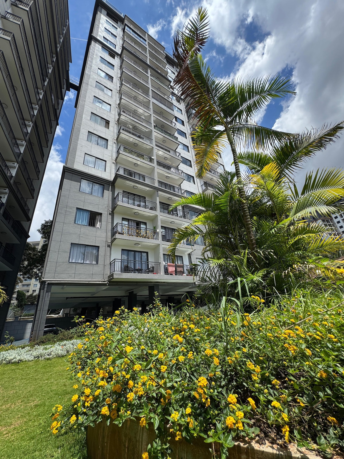 A tall modern apartment building is framed by vibrant green foliage and blooming yellow flowers at its base. The structure features multiple balconies with glass railings, allowing for natural light to brighten the living spaces. The sky above is dotted with clouds, creating a scenic backdrop.
