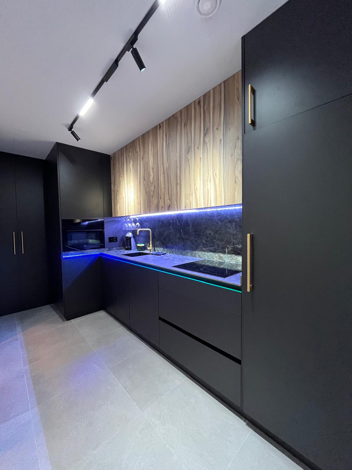 The kitchen features sleek, modern cabinetry in dark tones with wooden accents. A subtle blue LED light highlights the countertop area, which includes a sink and stovetop. Appliances, including a coffee maker, are integrated within the cabinetry, providing a cohesive and functional design.