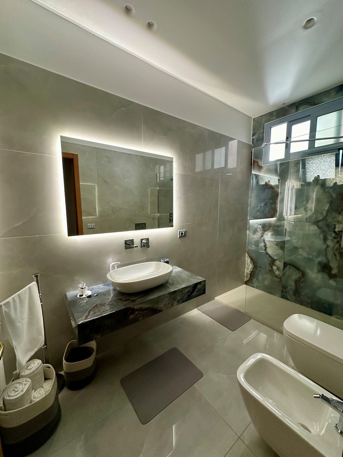A modern bathroom features sleek surfaces and a large backlit mirror above a minimalist sink. Elegant tile work decorates the shower area, and plush towels are neatly arranged beside the sink. Soft mats are placed on the floor for comfort.