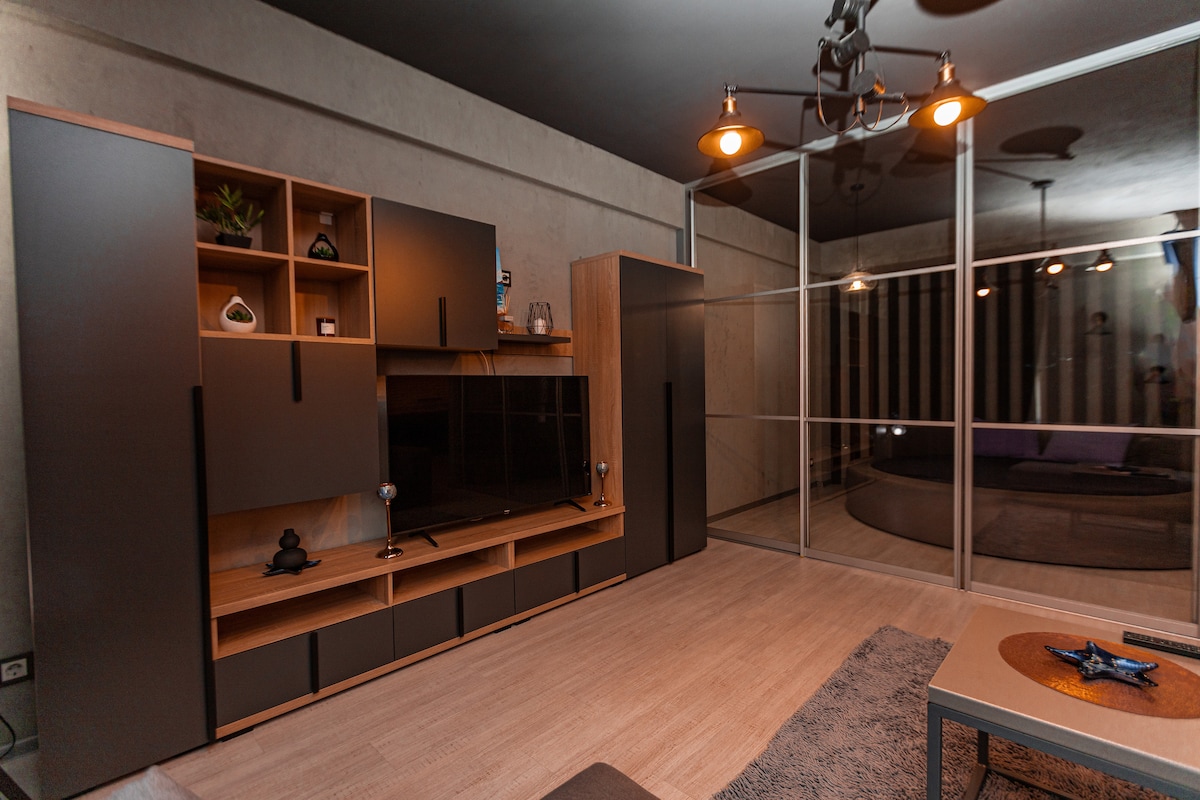 A modern living area is characterized by sleek cabinetry and a wall-mounted television. Natural light is permitted through large glass panels, creating an airy feeling. A comfortable sofa and a simple coffee table complete the space, enhanced by neutral tones and stylish lighting.