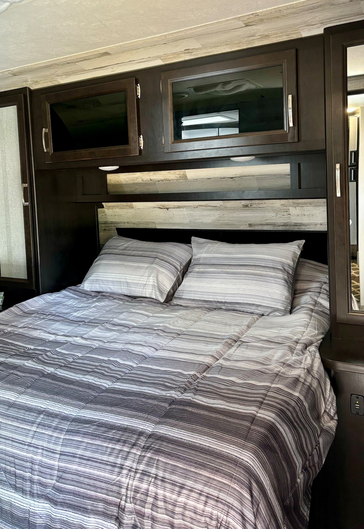 A cozy sleeping area is highlighted by a queen bed adorned with a striped gray and white comforter. The headboard features a subtle wood design, while dark cabinetry above provides additional storage. Soft lighting complements the relaxing space.