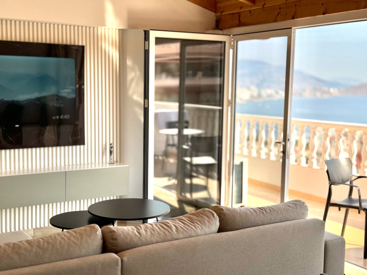 A modern living area features a comfortable sofa facing a large flat-screen TV. Floor-to-ceiling glass doors open to a private balcony with views of the distant mountains and shoreline. A sleek circular table is positioned on the polished floor, enhancing the open-air ambiance.