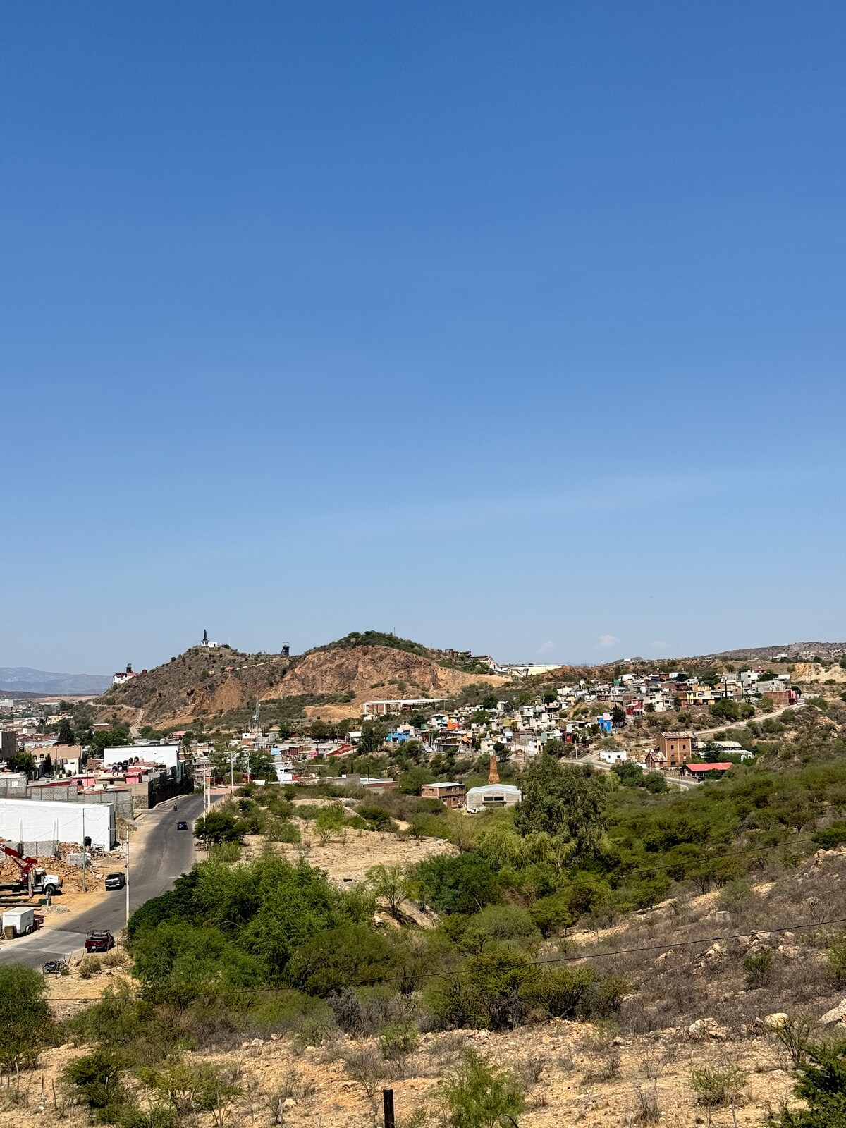 A wide view of a rural landscape is presented, featuring rolling hills in the background with structures dotting the hillside. The foreground reveals a mix of greenery and sparse vegetation, while a road leads through the scene, indicating easy access to the area.