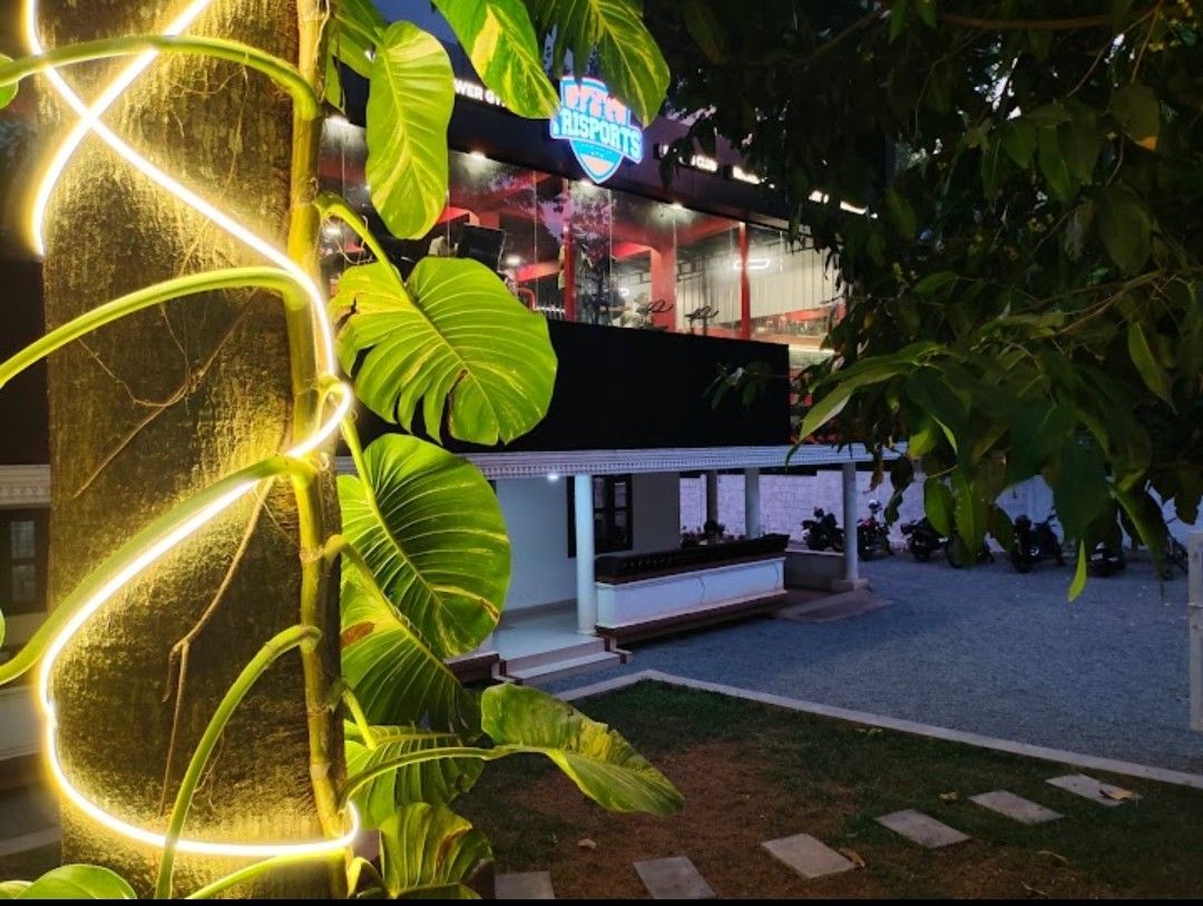 A well-lit exterior view of the facility is presented, showcasing a modern structure with large windows. Greenery decorates the foreground, with a bright vine wrapping around a tree trunk. A gravel area with parked vehicles adds to the accessible atmosphere.