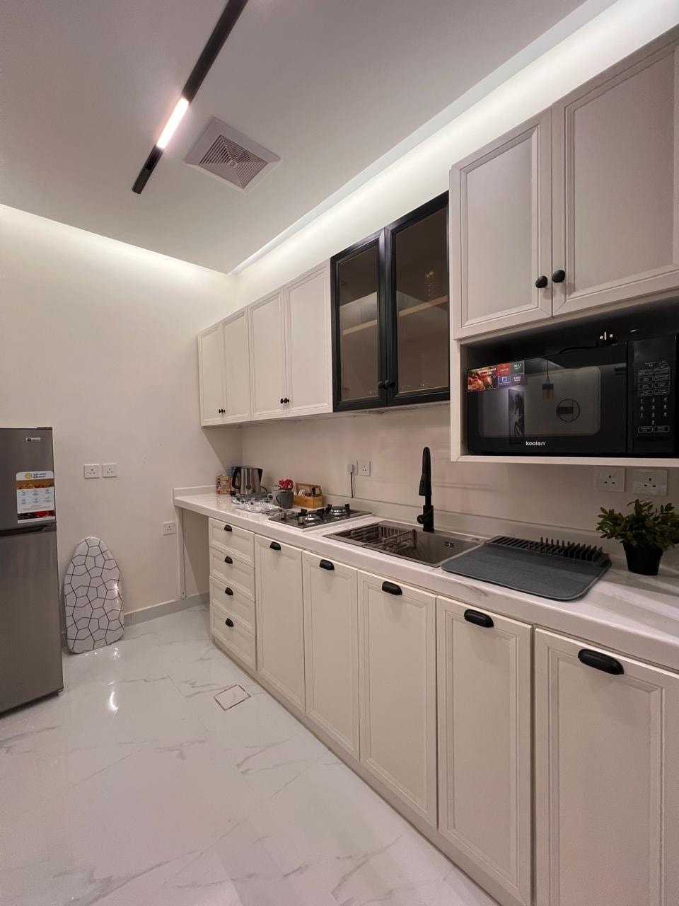 The kitchen features light-colored cabinetry with sleek black handles. A modern stainless steel refrigerator is positioned beside a microwave. A small sink and countertop area are visible, accompanied by essential kitchen appliances on the counter, all set against marble-effect flooring.