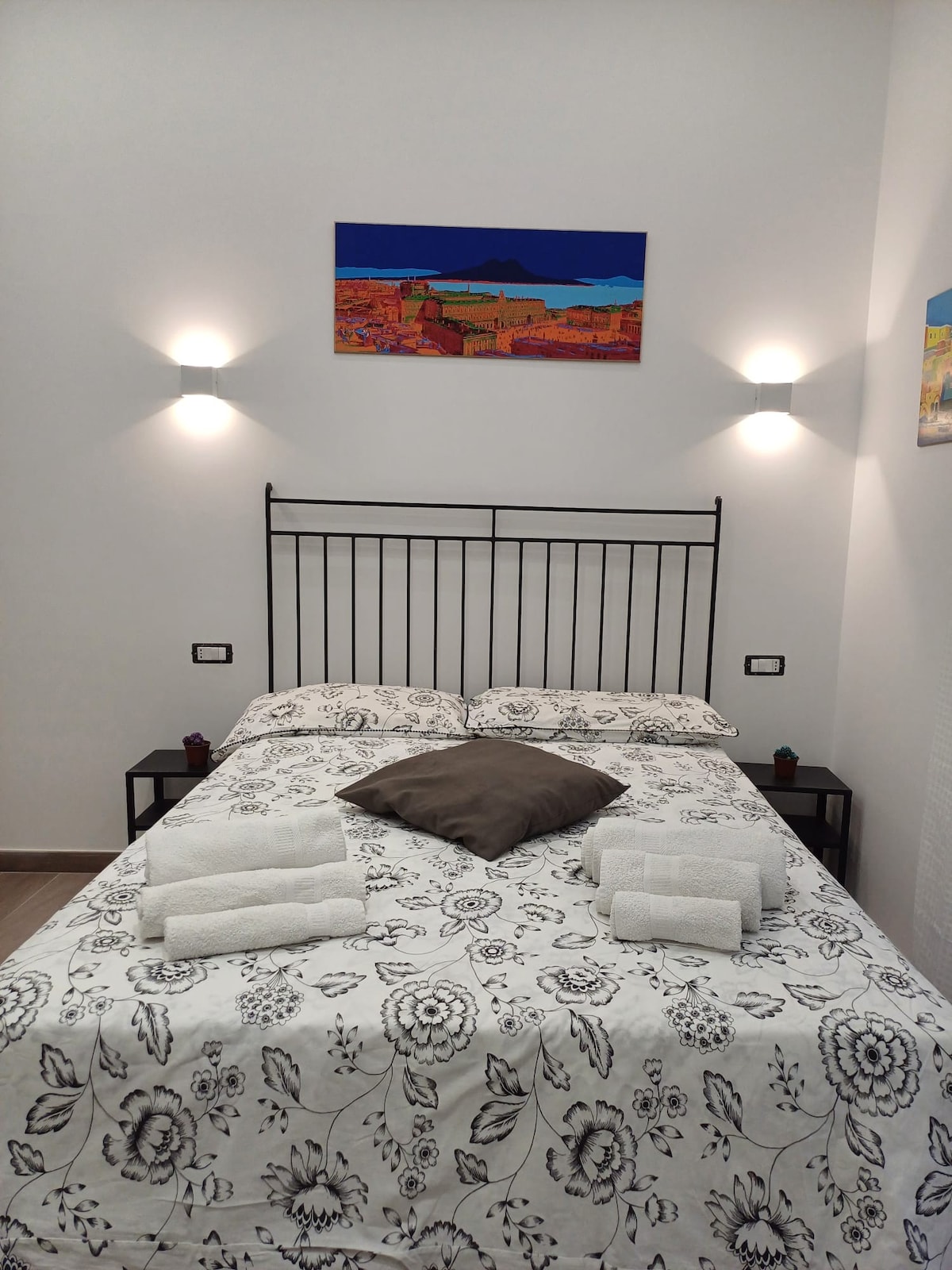 A double bed is centered in the room, adorned with a floral patterned duvet and a decorative brown pillow. Nightstands with small lamps are positioned on either side. Bright wall art and additional small artworks enhance the ambiance, while two towels are neatly arranged on the bed.