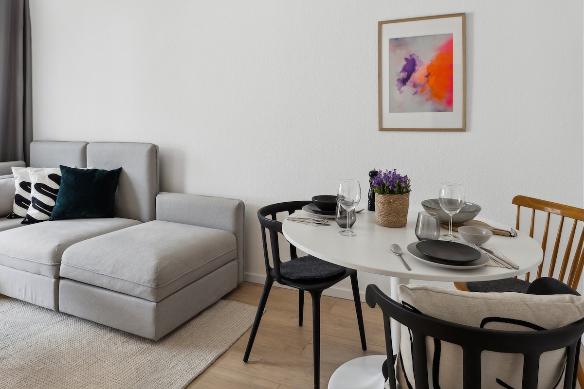 A modern living space features a comfortable gray sofa alongside a round dining table set for four. A colorful artwork adorns the wall, while a small vase with purple flowers adds a touch of freshness. Natural light brightens the room, enhancing its inviting feel.