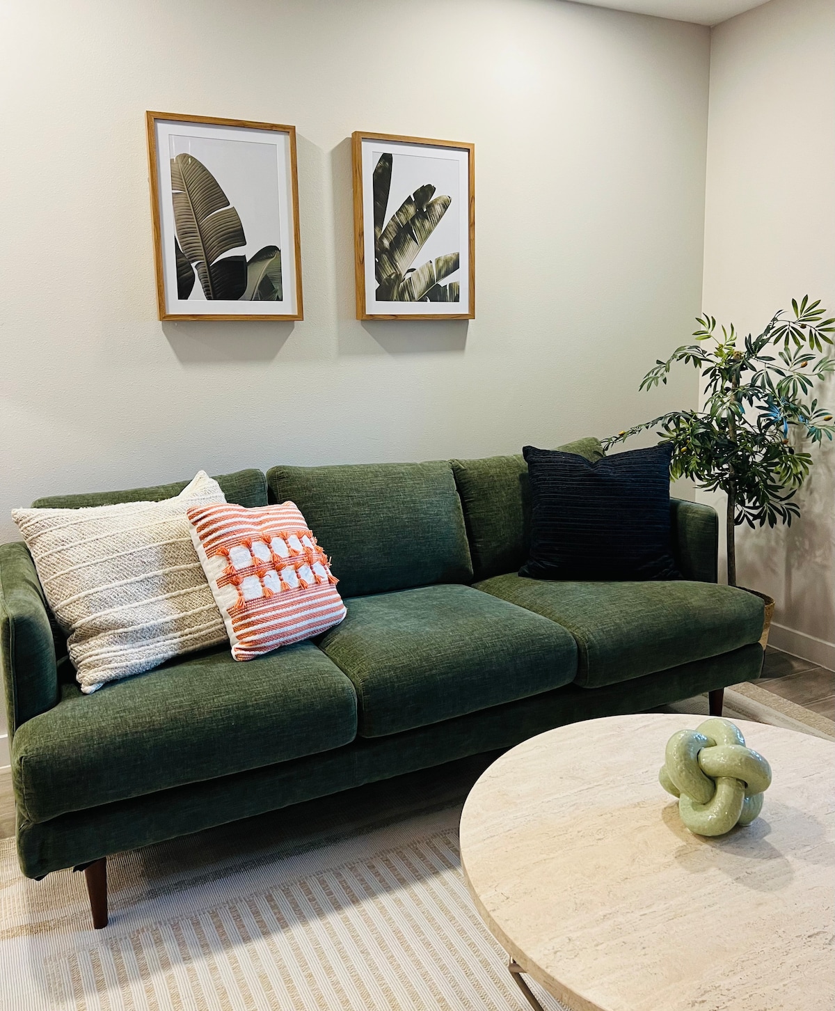 A cozy seating area is furnished with a green couch, accentuated by patterned pillows in various colors. Above, two framed artworks featuring leaves add a natural touch. A round coffee table made of light-colored material is positioned within the space, complemented by a decorative plant.