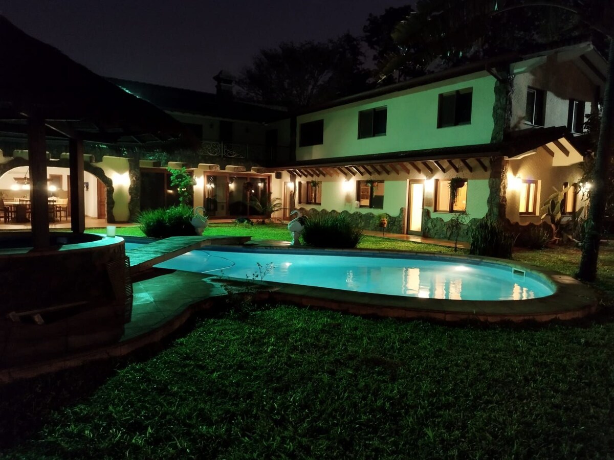 A tranquil night scene features a private pool illuminated by soft lighting, surrounded by lush green grass. The villa showcases a two-story structure with outdoor seating under a gazebo and large windows allowing a view of the inviting interior.