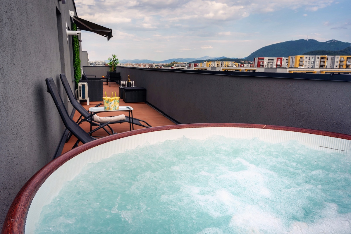 A private terrace features a hot tub surrounded by wooden decking, with two lounge chairs positioned nearby. The skyline and distant mountains are visible in the background, creating a serene outdoor space for relaxation.