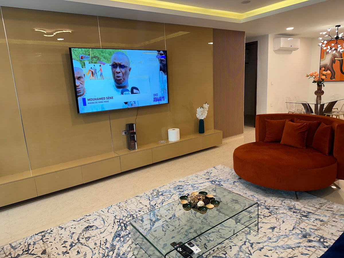 A modern living area features a large round orange sofa and a glass coffee table with a decorative arrangement of items. A flat-screen TV is mounted on the wall, flanked by sleek cabinetry and softly illuminated by ambient lighting.