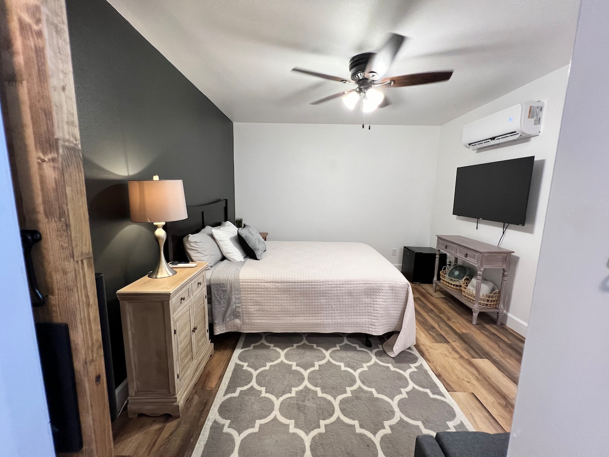A well-appointed room features a spacious bed dressed in neutral tones. A bedside lamp provides soft lighting. A television is mounted on the wall opposite the bed. A patterned area rug plays contrast with the wooden floor. An air conditioning unit is also visible.