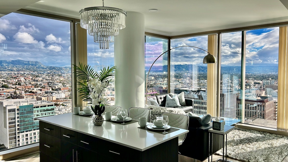 A spacious living area is highlighted by floor-to-ceiling windows offering expansive views of the Los Angeles skyline and surrounding mountains. A modern chandelier hangs above a dark dining table set for two, while a contemporary sofa is positioned in front of the view, accented with a floor lamp and decorative plants.