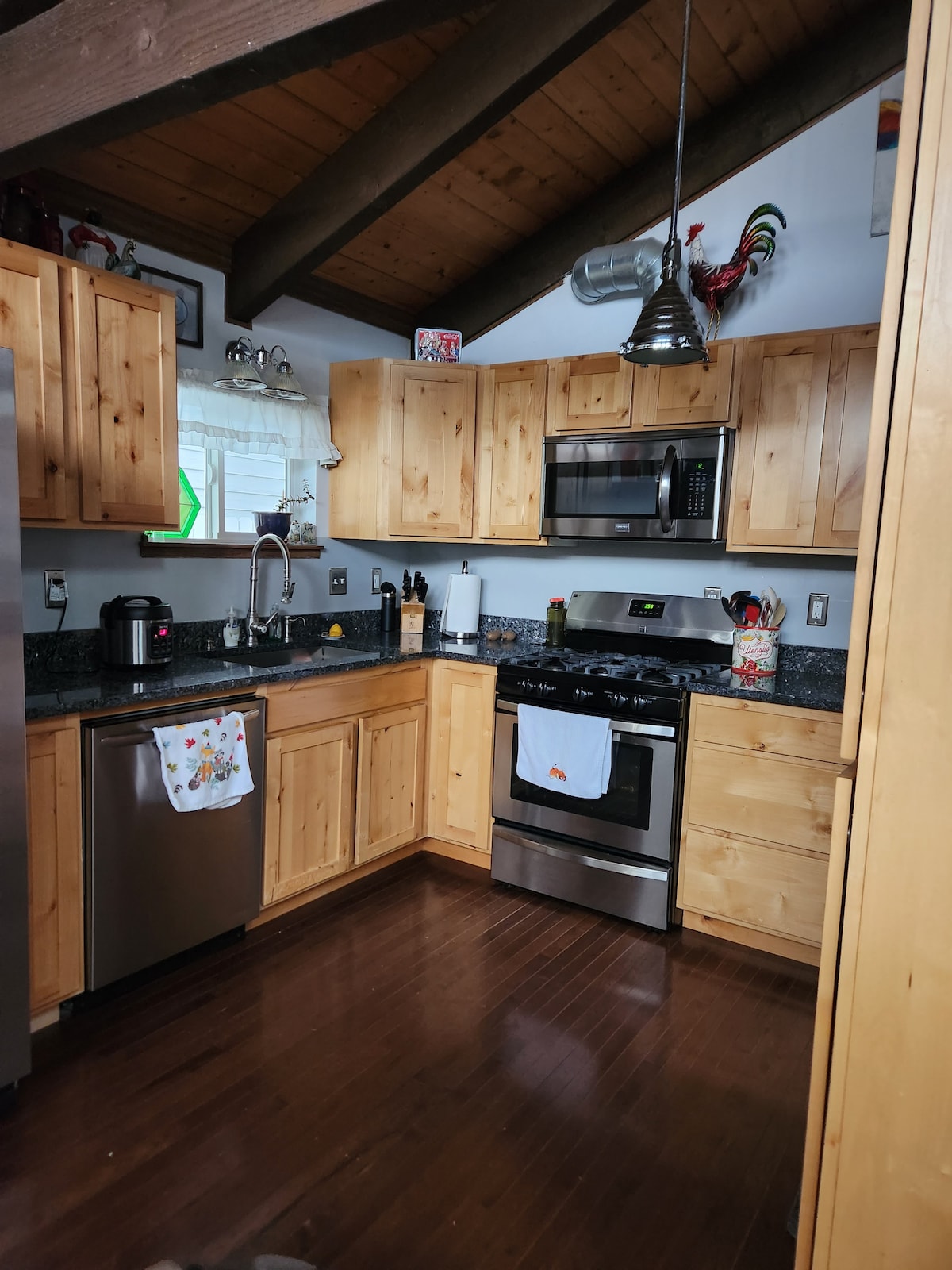 A kitchen featuring wooden cabinetry and dark granite countertops is illuminated by natural light. Stainless steel appliances, including a refrigerator and stove, are present. A hanging light fixture adorns the ceiling, and decorative accents can be seen on the shelves.