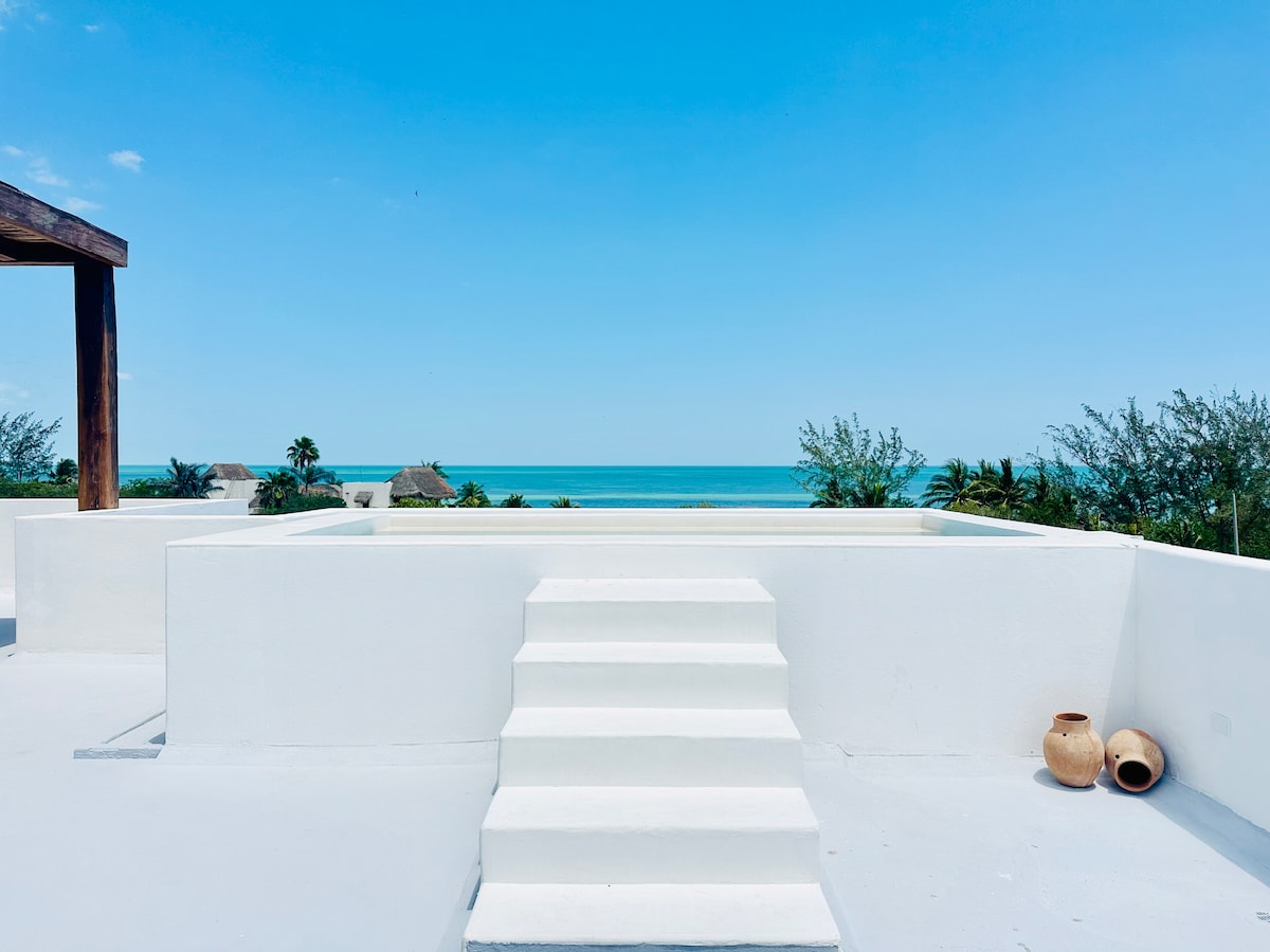 A sunlit terrace features a white walled platform with steps leading down to a beach view. Lush greenery and a glimpse of the ocean are visible in the background, enhancing the serene outdoor space.