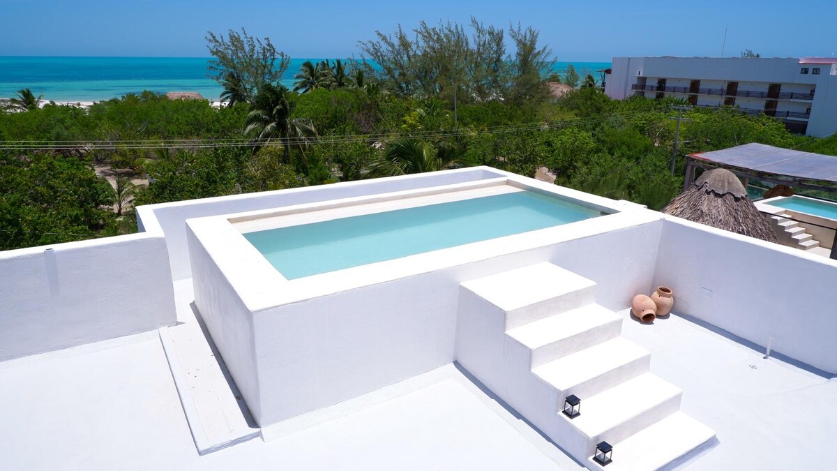 A rooftop pool is showcased, featuring clear water and surrounded by white surfaces. Steps lead down from the pool area, offering views of the lush greenery and distant ocean. The serene setting highlights the tranquility of the surroundings and provides an inviting space for relaxation.