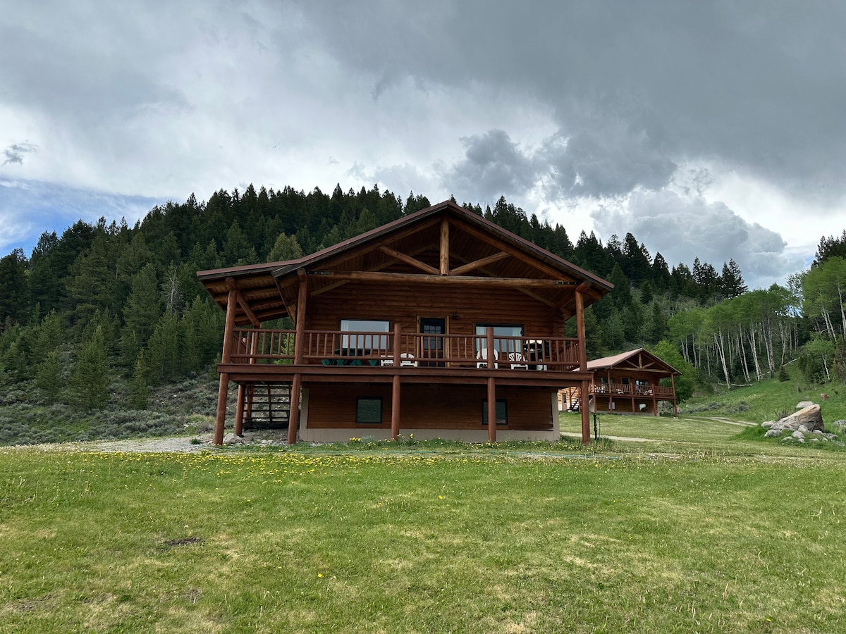 A split-level log lodge is set against a backdrop of dense greenery, with expansive decks on both levels. The structure features large windows allowing views of the surrounding natural landscape, and grassy areas dotted with yellow wildflowers enhance the peaceful ambiance.