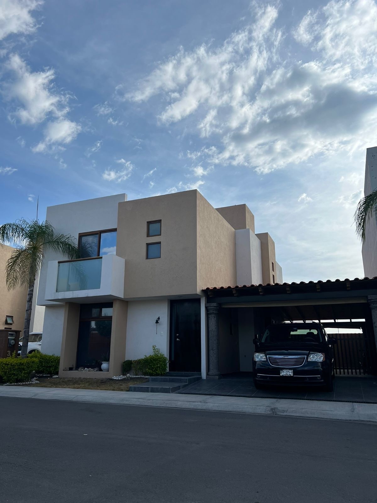 The contemporary two-story building features a mix of cream and light brown exterior walls. Large windows allow natural light to fill the space, while a covered carport accommodates vehicles. Lush greenery is present alongside the entrance, contributing to the welcoming appearance.