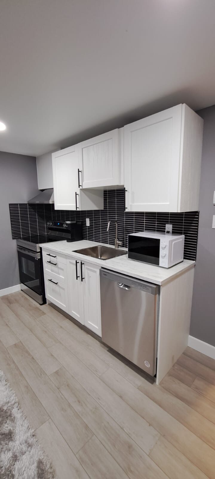 A modern kitchen is equipped with essential appliances, including an oven, microwave, and dishwasher. Sleek white cabinetry contrasts with dark tile backsplash, while a stainless-steel sink is positioned beneath overhead storage. Light hardwood flooring adds warmth to the space.