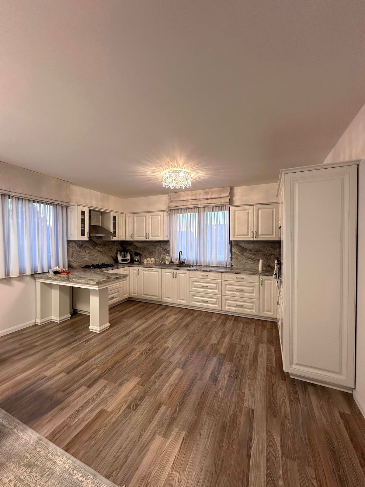 A modern kitchen features white cabinetry and a marble backsplash, illuminated by a stylish chandelier. Natural light enters through large windows, highlighting the spacious layout. Appliances, including a stove and coffee maker, are neatly arranged to enhance functionality.