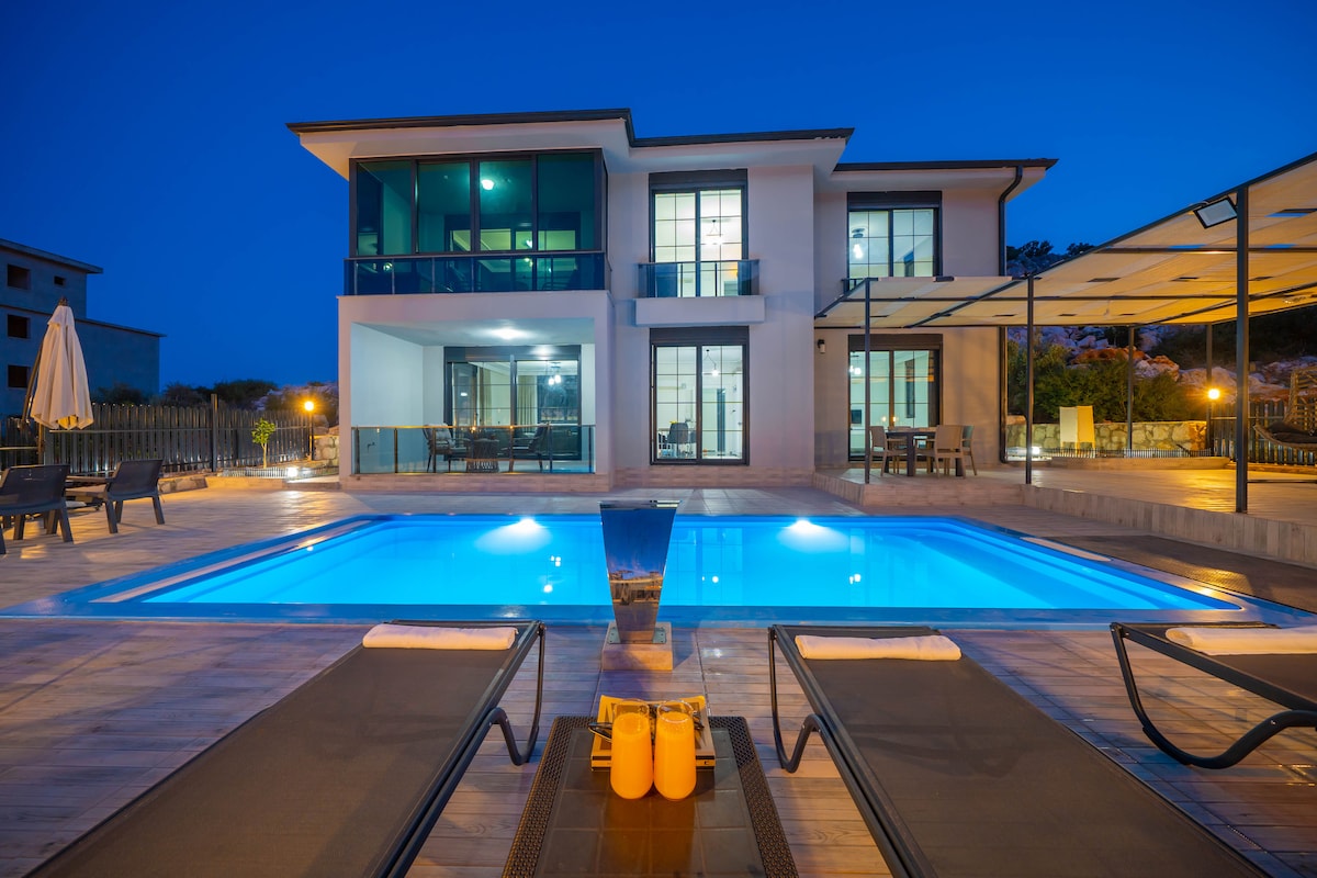 The modern villa is highlighted by a geometric pool illuminated in blue tones, surrounded by a spacious wooden deck. Loungers are positioned nearby, and large glass doors reveal the interior. Soft lighting enhances the exterior, showcasing the villa's contemporary architecture.