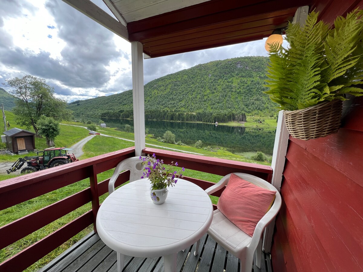 A small outdoor seating area features a round white table with a potted flower and two chairs. The view overlooks a serene lake, surrounded by lush green hills, with a tractor visible on the path. The area is shaded by a covered balcony.
