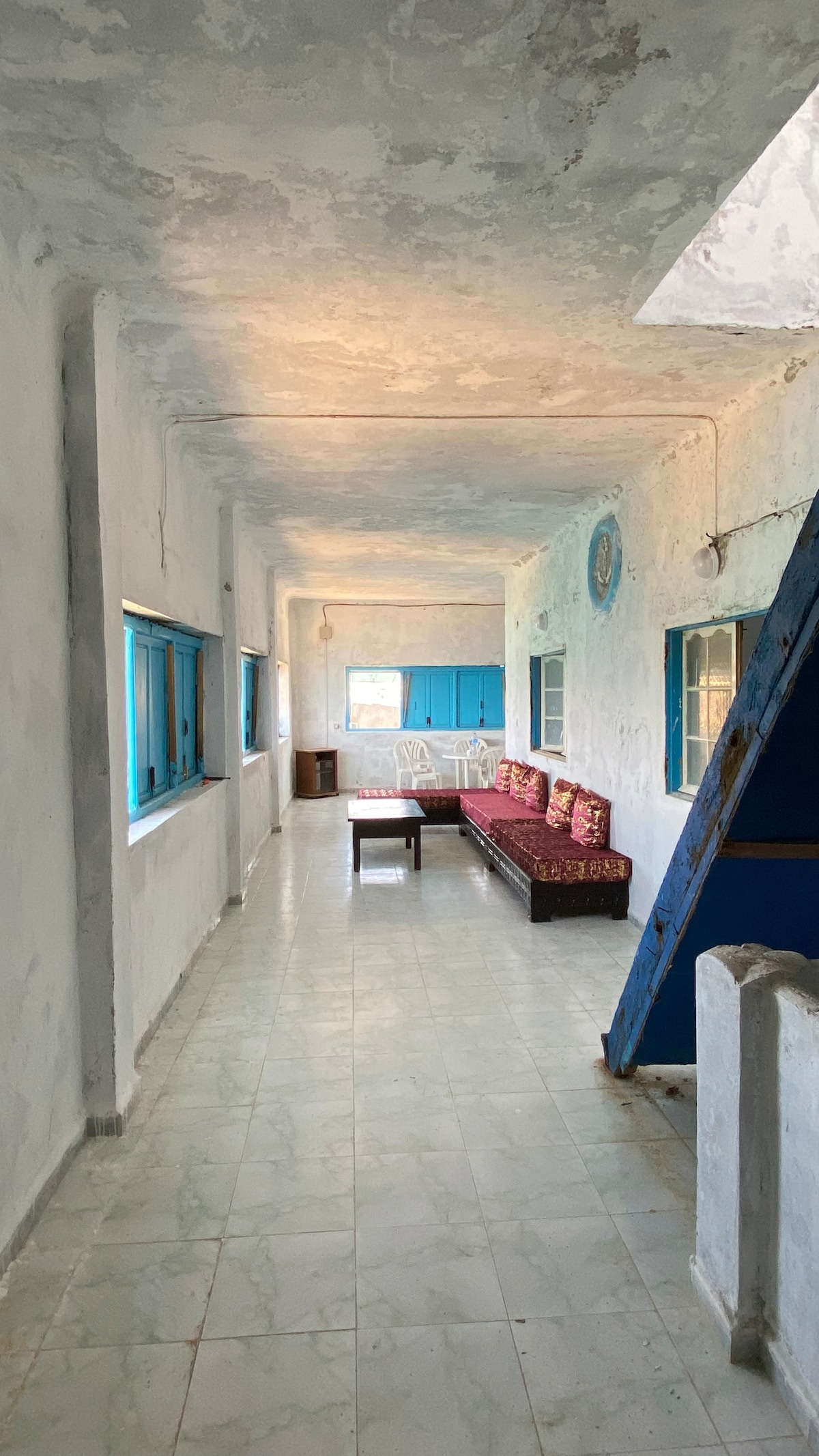 A long, airy hallway features a whitewashed wall with blue accents. Natural light floods in through multiple windows, illuminating a cozy seating area with patterned cushions. A wooden table is positioned nearby, while a staircase leads upwards, adding depth to the space.