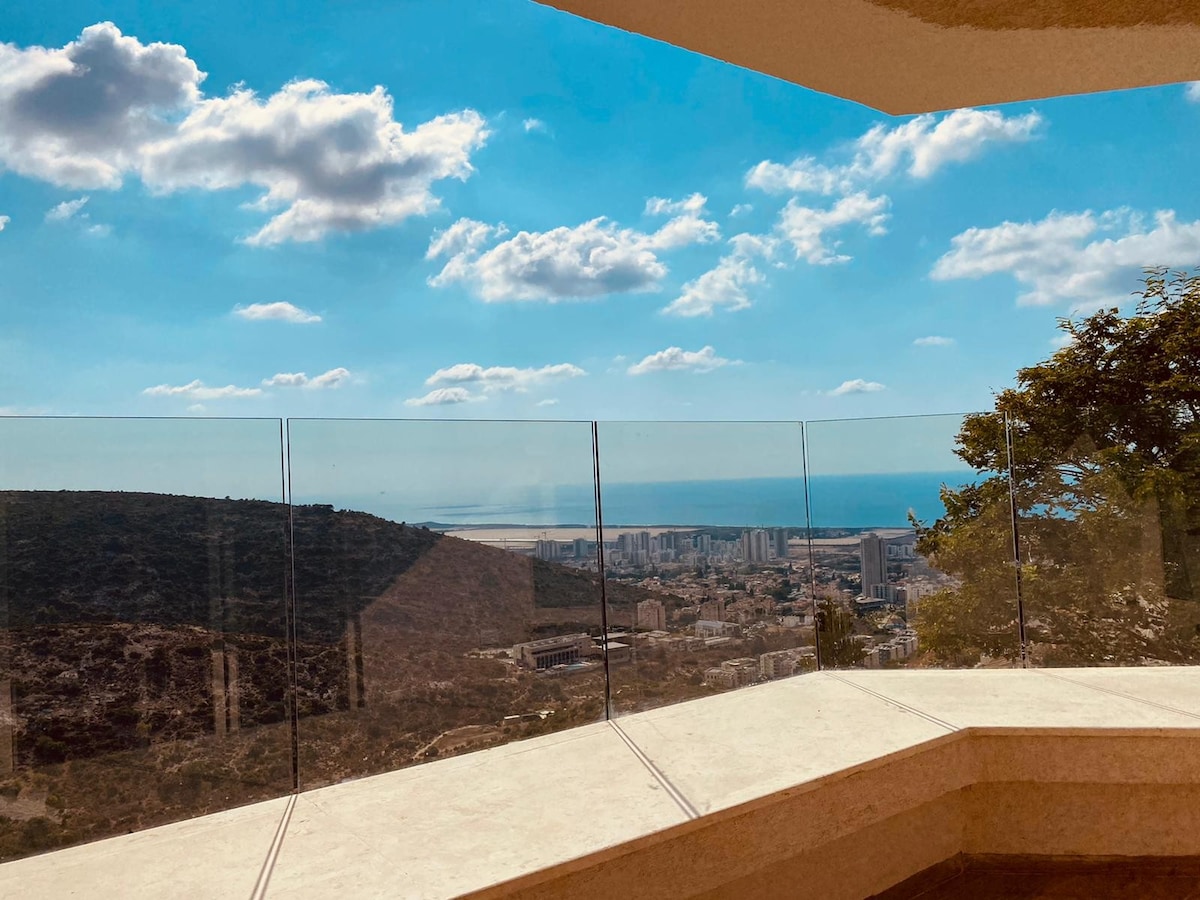 A spacious terrace features a panoramic view of the coastline and cityscape, framed by clear glass railings. The sky is sunny with scattered clouds, and the surrounding landscape includes hills and greenery.