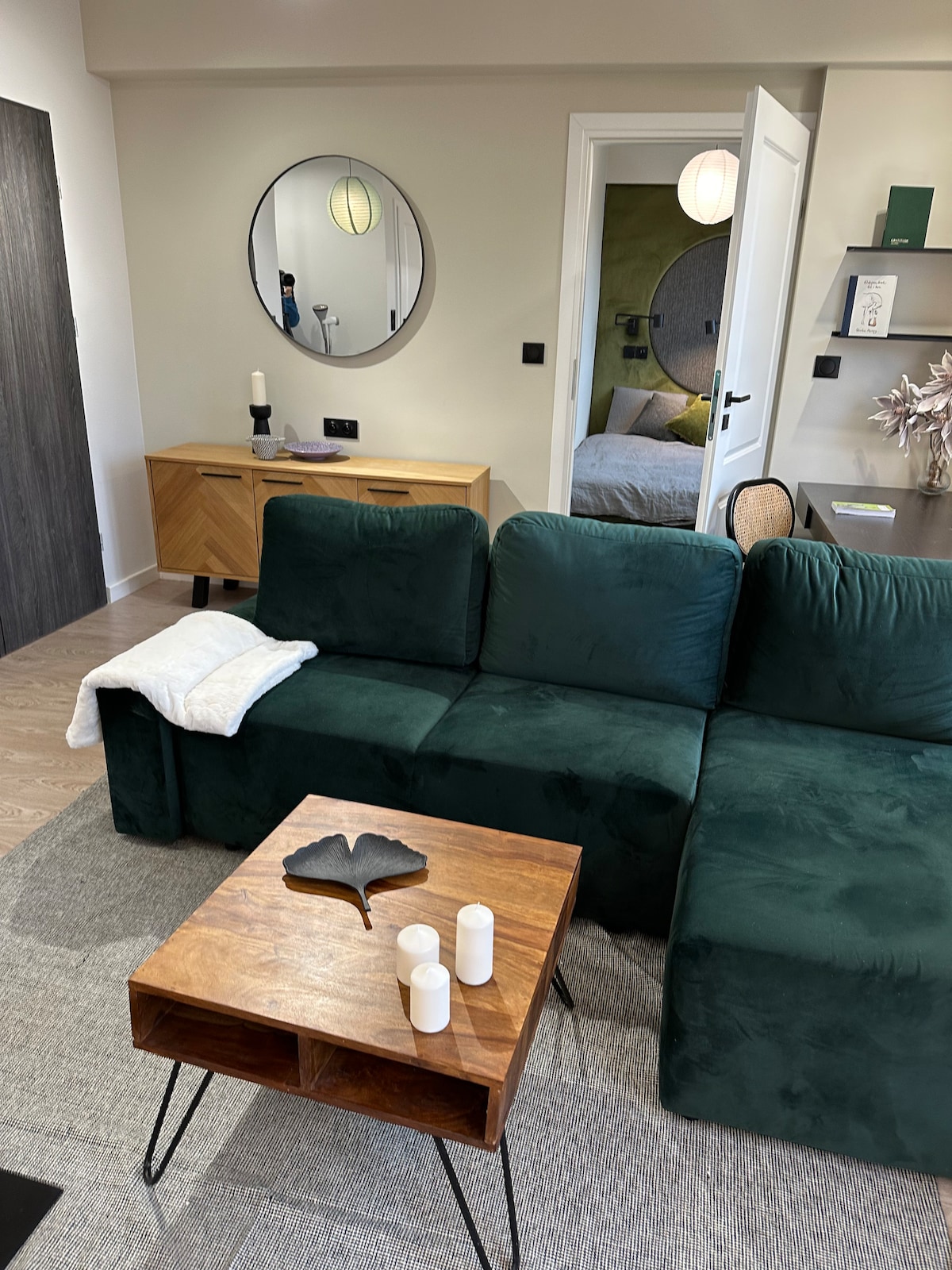 A comfortable living area features a green sofa positioned around a wooden coffee table. Candles are arranged decoratively on the table, which has a natural finish. A round mirror hangs on the wall, reflecting the cozy ambiance of the connected space.