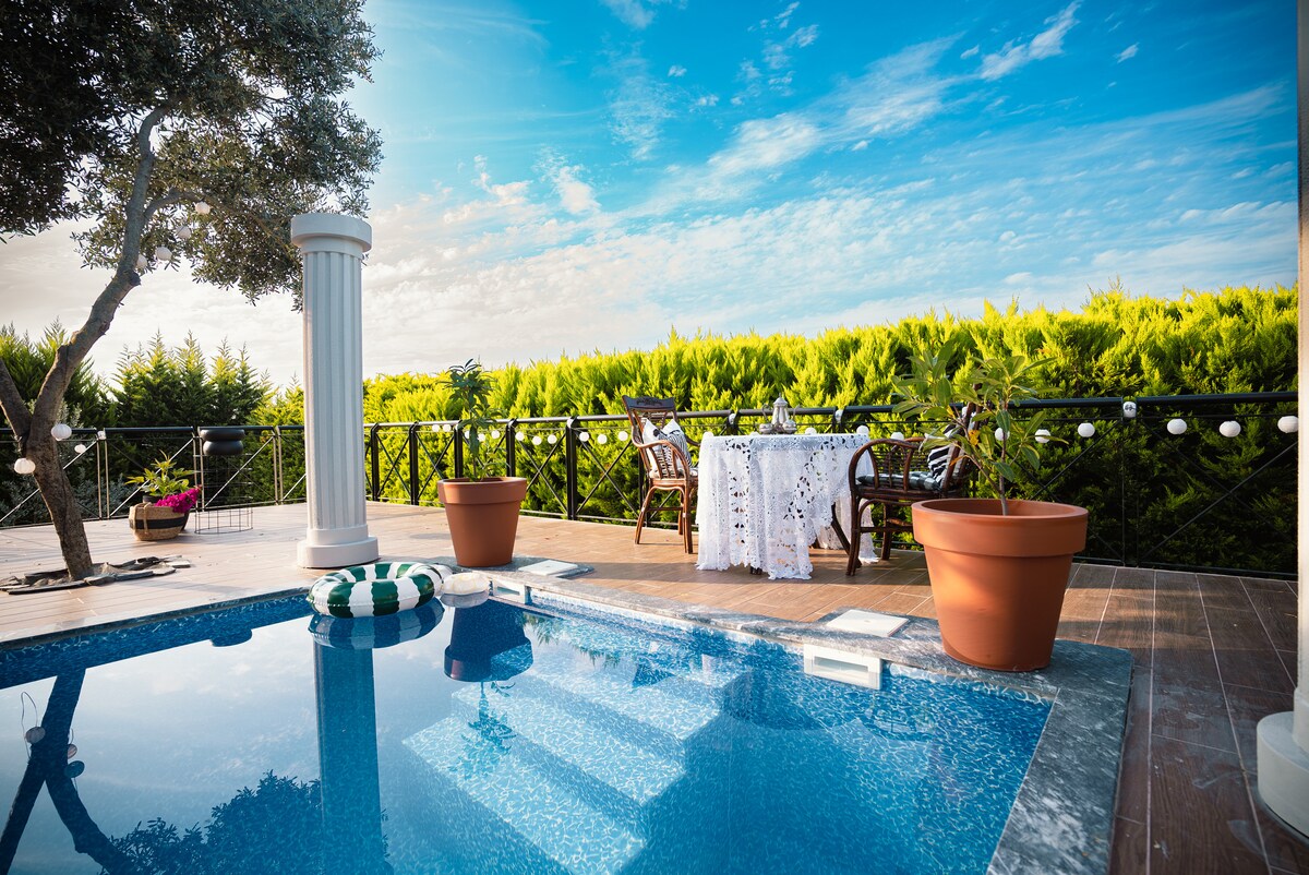 A tranquil outdoor setting features a swimming pool adorned with a colorful float. A dining area is arranged on a deck, with a table dressed in white linens and surrounded by wooden chairs. Lush greenery forms a serene backdrop under a blue sky.