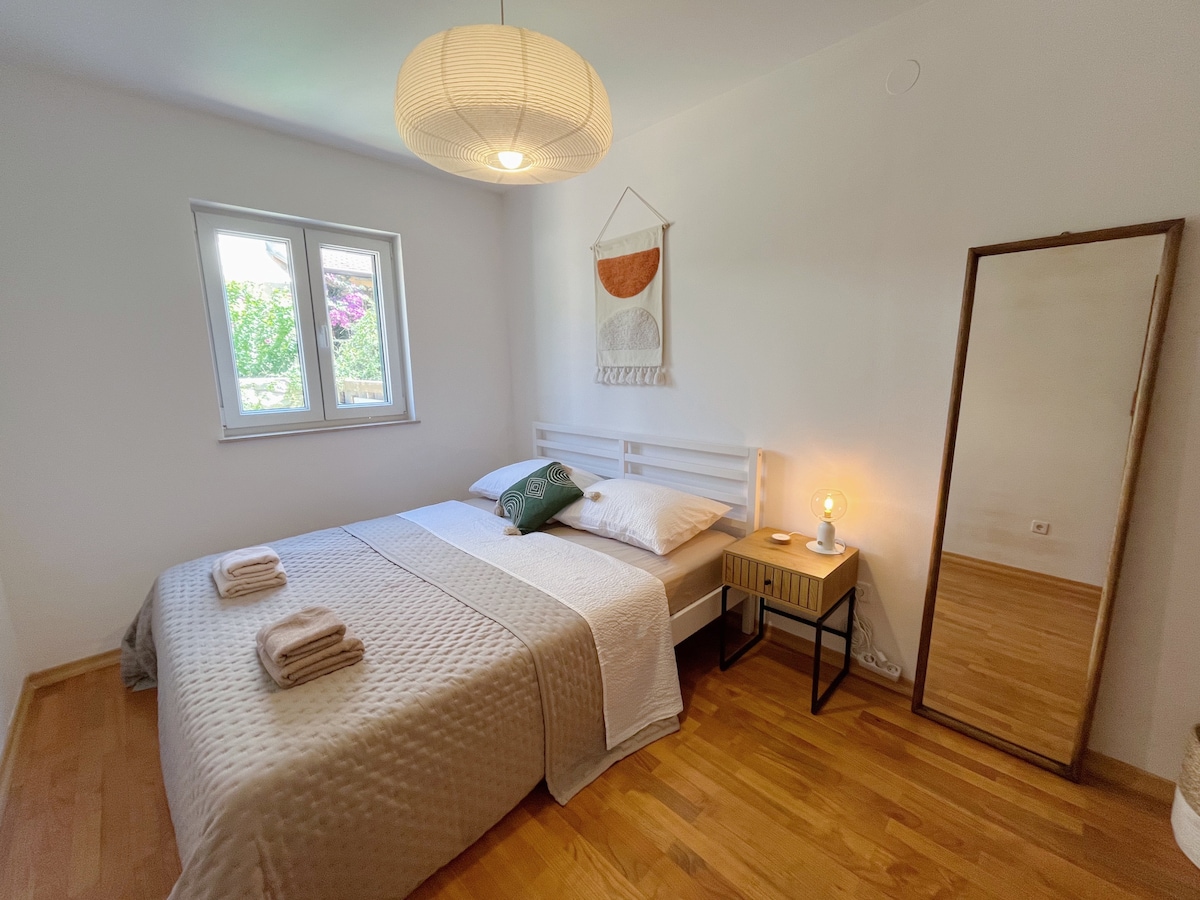 A serene bedroom is presented with a queen-sized bed dressed in light linens. A decorative wall hanging and soft lamp enhance the ambiance. A full-length mirror reflects the neutral decor, while natural light enters through two windows framed by greenery.
