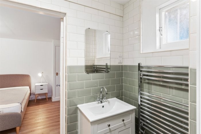 A modern bathroom with green tile accents features a sleek white vanity topped with a stylish sink. A rectangular mirror is mounted above the sink, and a heated towel rail is situated on the adjacent wall. Natural light enters through a small window.