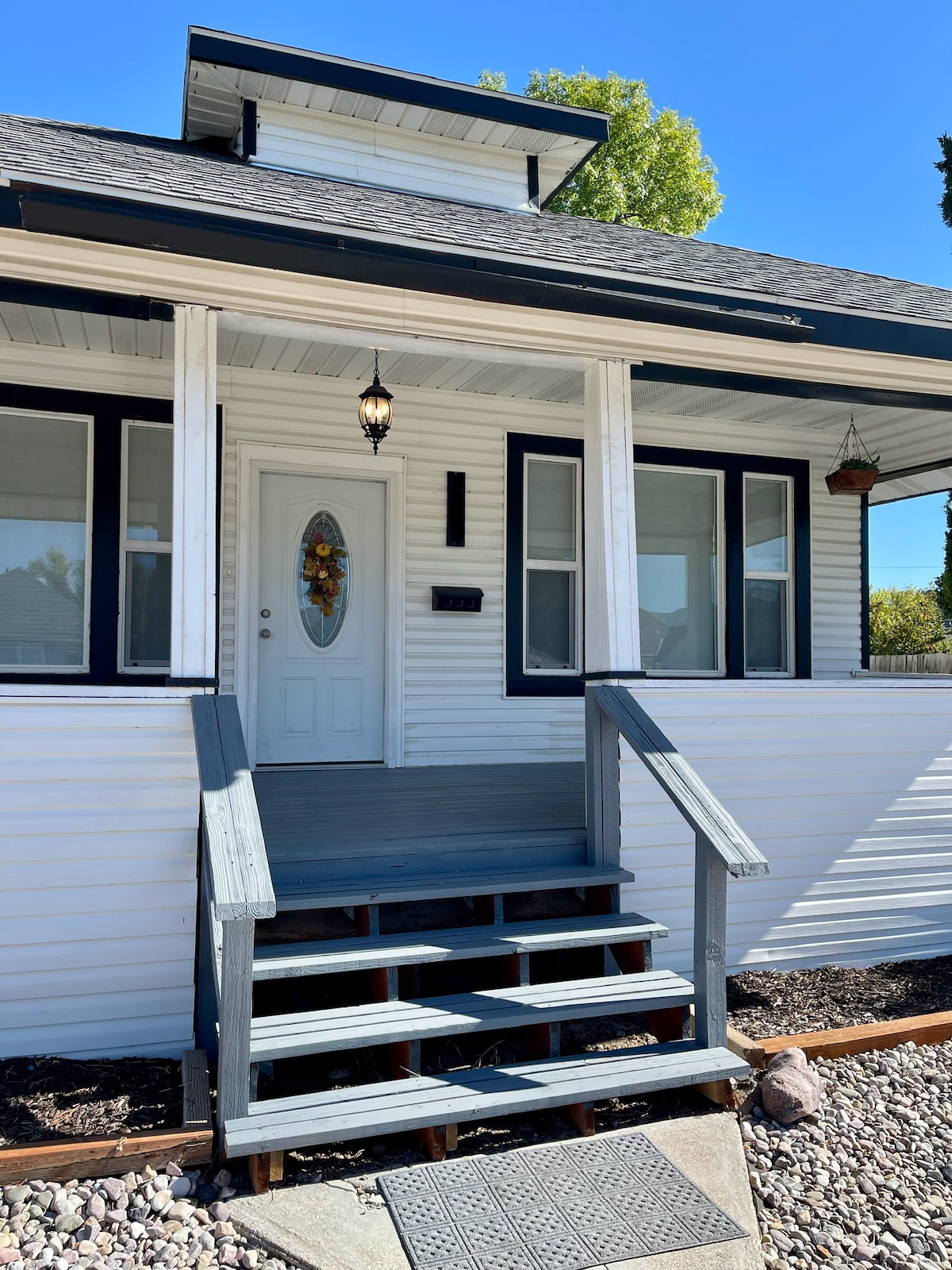 The front entrance of the home features a white door framed by glass panels, accented by a decorative wreath. A small porch is accessed via a set of wooden steps, with a light fixture hanging above the entryway.