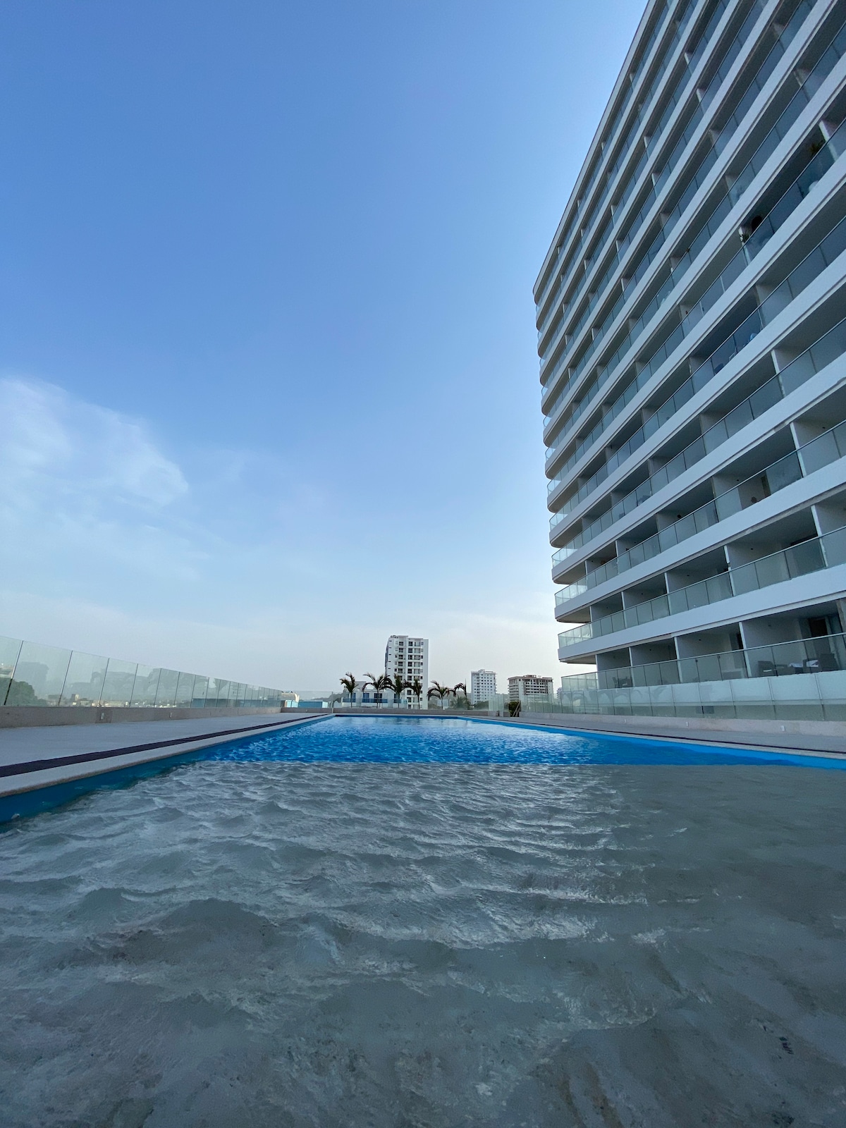 A rooftop swimming pool is visible, bordered by a glass railing. The modern building facade towers beside the pool, reflecting a clear blue sky. Palm trees can be seen in the distance, adding to the serene atmosphere.