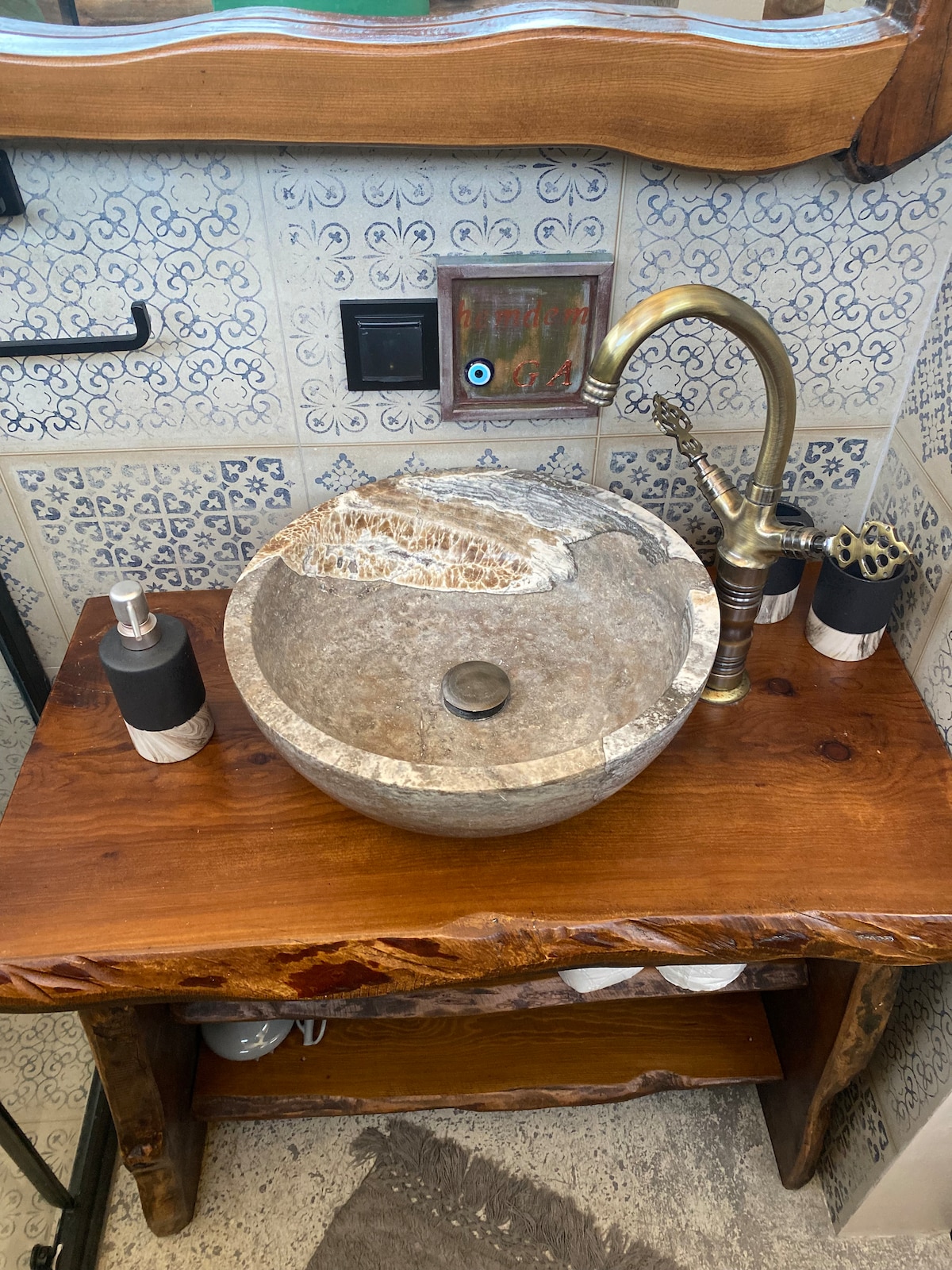 A stone sink is set on a wooden vanity, featuring rustic finishes. The sink's natural texture is complemented by stylish soap dispensers and toothbrush holders. A decorative backsplash with intricate blue tiles enhances the overall aesthetic of the space.
