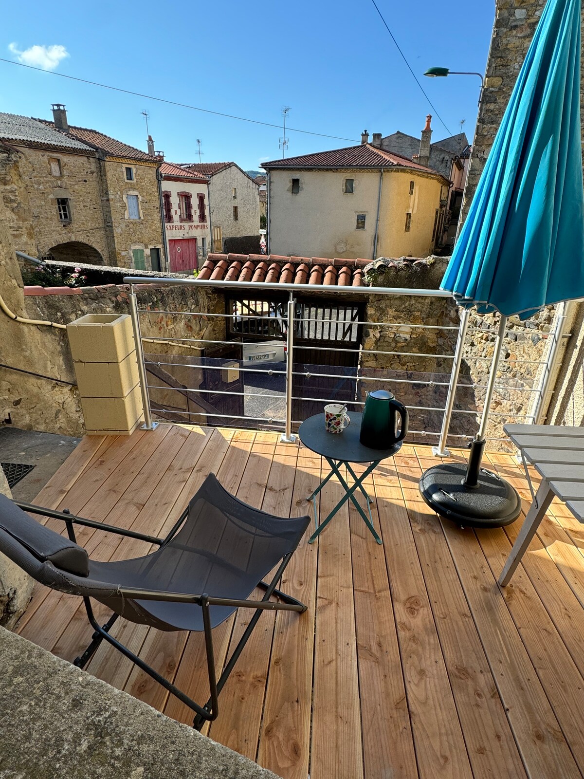 An outdoor terrace features wooden decking with a small round table and a chair. A blue umbrella offers shade, and a kettle is placed on the table. The view includes neighboring buildings with tiled roofs, and a stone wall provides a rustic backdrop.