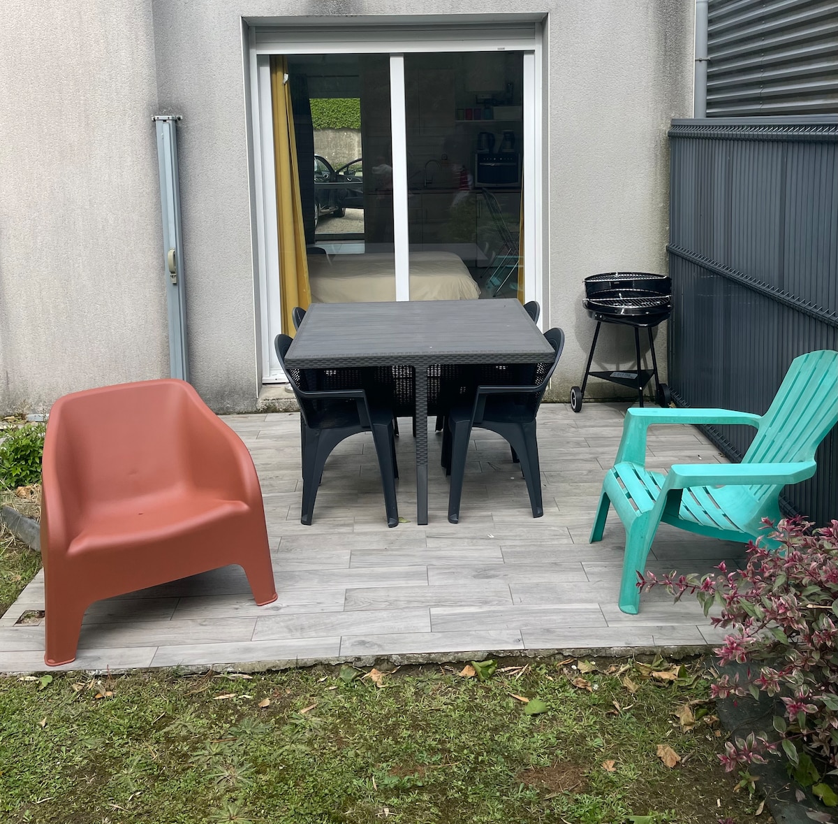 An outdoor terrace area is depicted, featuring a dark table accompanied by several chairs in different colors. A grill is placed nearby, and lush greenery borders the space. Large glass doors provide access to the interior, which is partially visible.