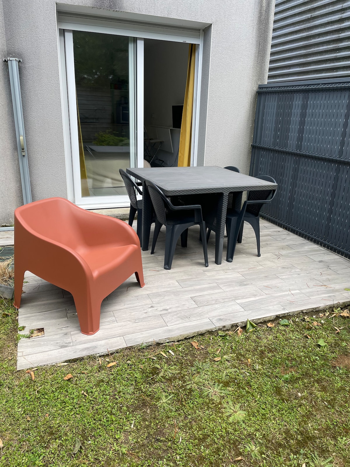 A small terrace area features a rectangular table surrounded by six black chairs. One orange chair is positioned nearby, adding a pop of color. The outdoor space is bordered by grass and has large glass doors leading inside, allowing natural light to flow through.